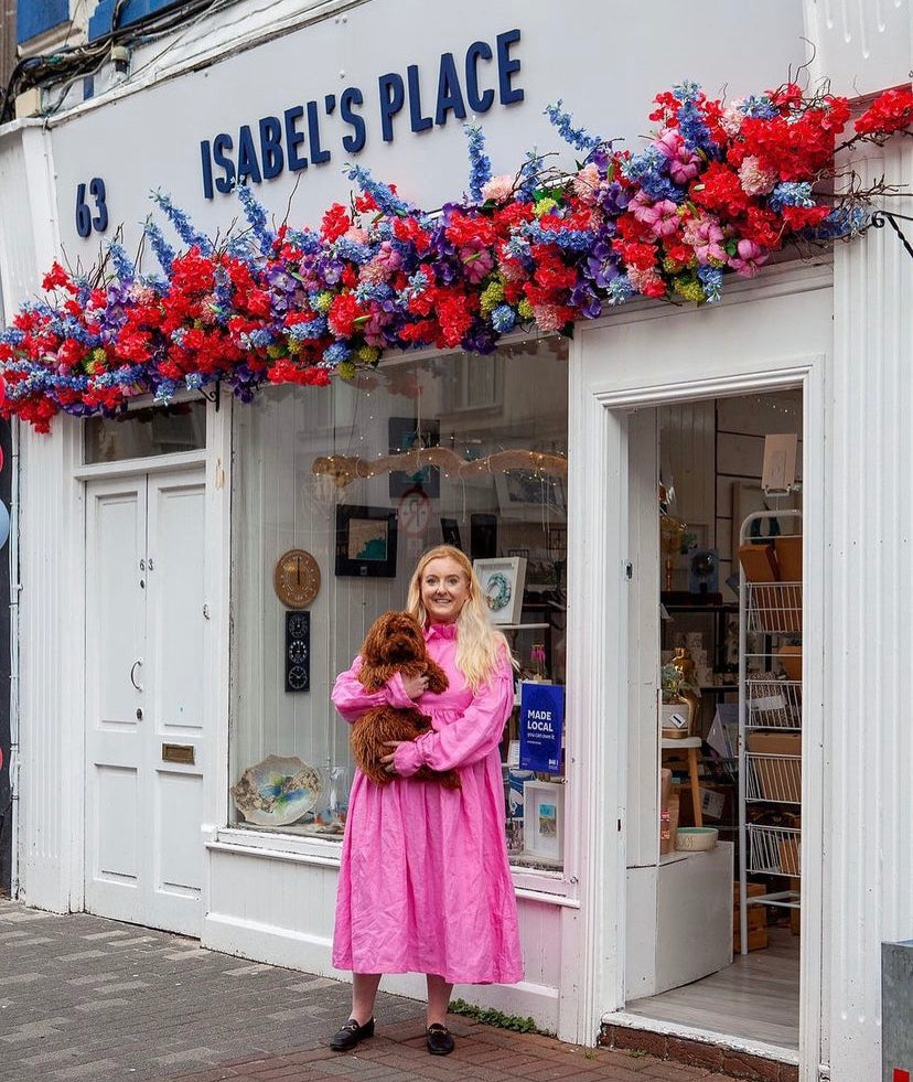 Woman in a pink dress holding a small brown dog outside Isabel's Place, with red and blue flowers