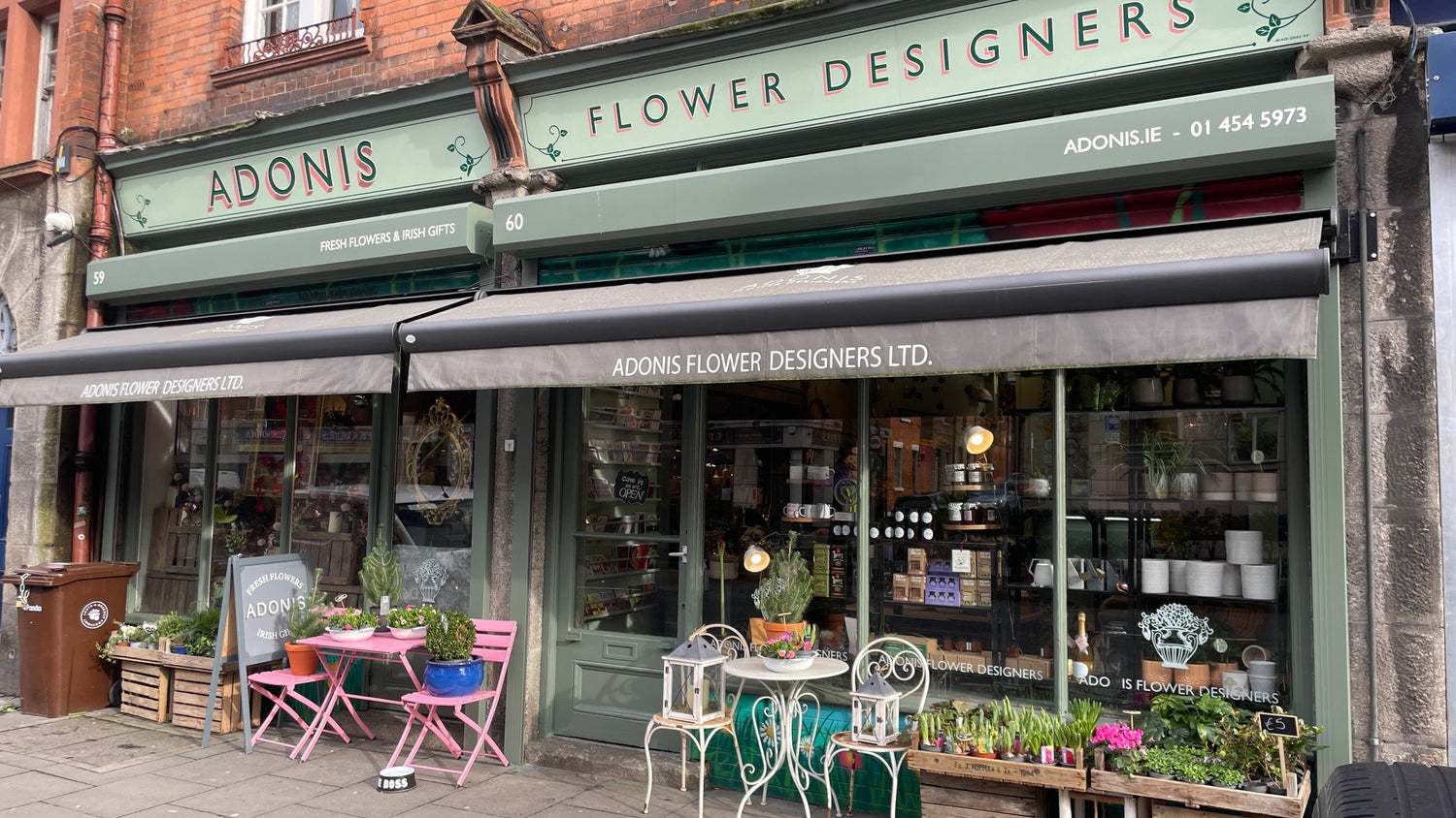 Flower shop frontage with green and white signage, awnings, and pink chairs outside a brick building