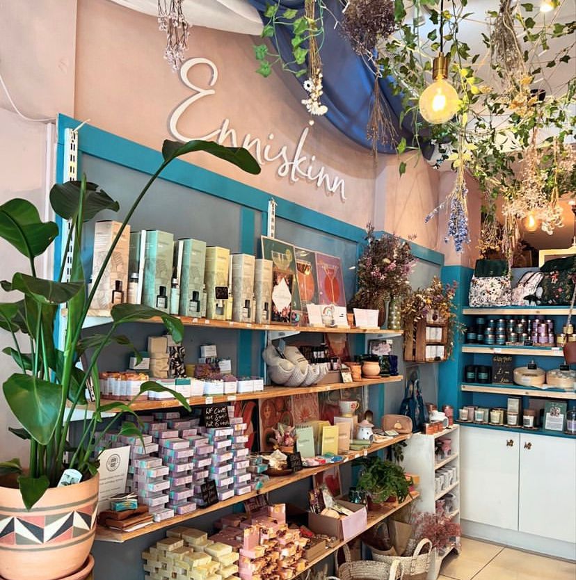 A shop display with shelves of candles and skincare jars, blue trim, hanging lights, and greenery